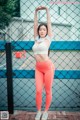 A woman in a white top and orange leggings posing in front of a fence.